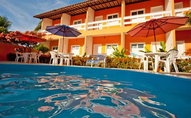 Piscina azul em primeiro plano com prédio de dois andares de fachada laranja ao fundo, com mesas e guarda-sóis vermelhos.