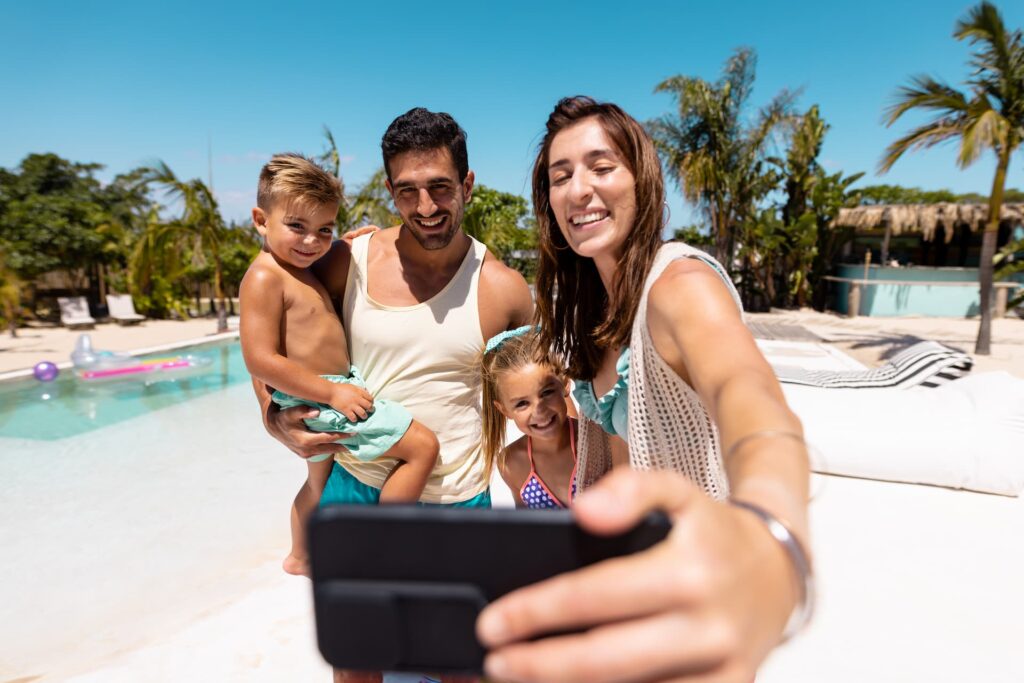Família biracial sorridente tirando uma selfie com celular à beira de uma piscina ensolarada.