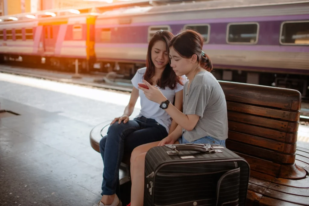 Grupo de amigos sentados em um banco de madeira em uma estação de trem, conversando e usando smartphones enquanto aguardam o embarque.