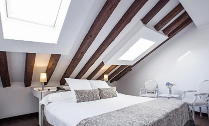 Suíte com teto inclinado e vigas de madeira aparentes, equipada com cama de casal grande e janelas de sótão em madri.