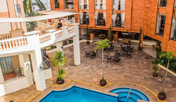 Pátio interno com piscina azul, mesas de café e prédios de cor terracota com varandas clássicas.