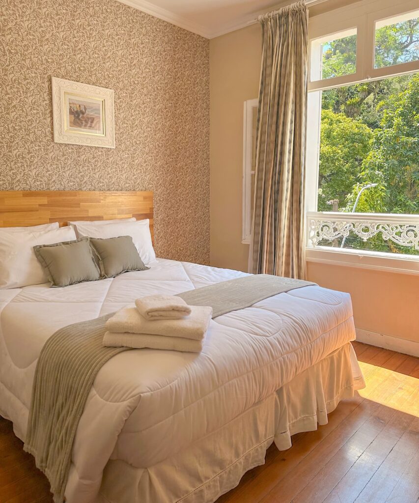 Quarto charmoso com cama de casal, enxoval branco, parede com papel de parede clássico e uma janela com vista para árvores verdes. Feriados Roteiros Curtos