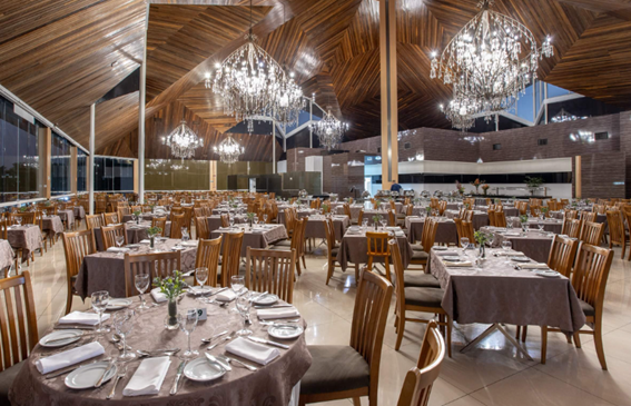 Salão de restaurante amplo e luxuoso, com mesas montadas, cadeiras de madeira e grandes lustres de cristal sob um teto inclinado de madeira.