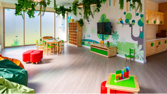 Sala colorida com mesas pequenas, cadeiras, pufes, prateleiras de brinquedos e decoração de floresta nas paredes.