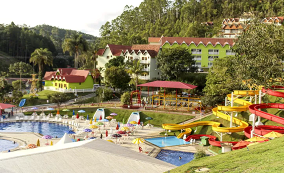 Parque aquático com toboáguas coloridos (amarelo e vermelho) e piscinas, com prédios de estilo europeu ao fundo em uma área montanhosa.