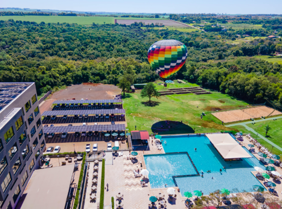 Vista aérea mostrando uma piscina grande, prédios do resort, estacionamento com painéis solares e um balão de ar quente colorido sobrevoando a área verde.