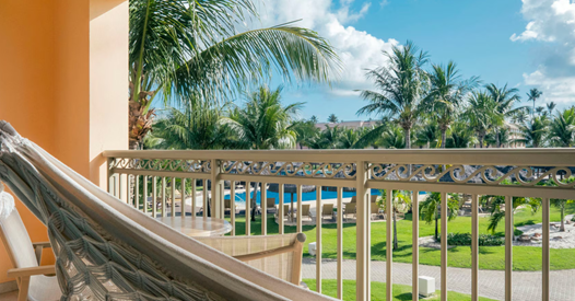 Vista a partir de uma varanda com rede, mostrando gramado verde, coqueiros e uma piscina ao fundo.